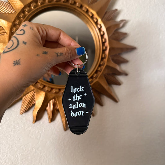 Hand holding a keychain with 'lock the salon door' text in front of a decorative mirror.