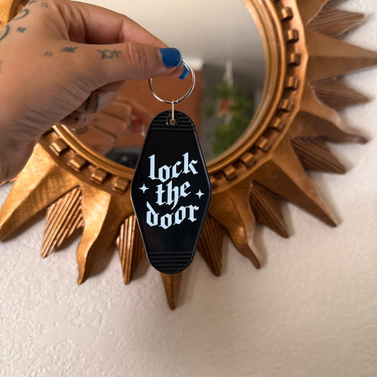 Keychain with 'lock the door' text held by a hand in front of a decorative mirror.