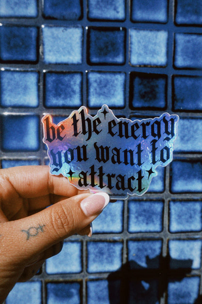 Hand holding a colorful sticker with 'be the energy you want to attract' text against a blue tiled background.