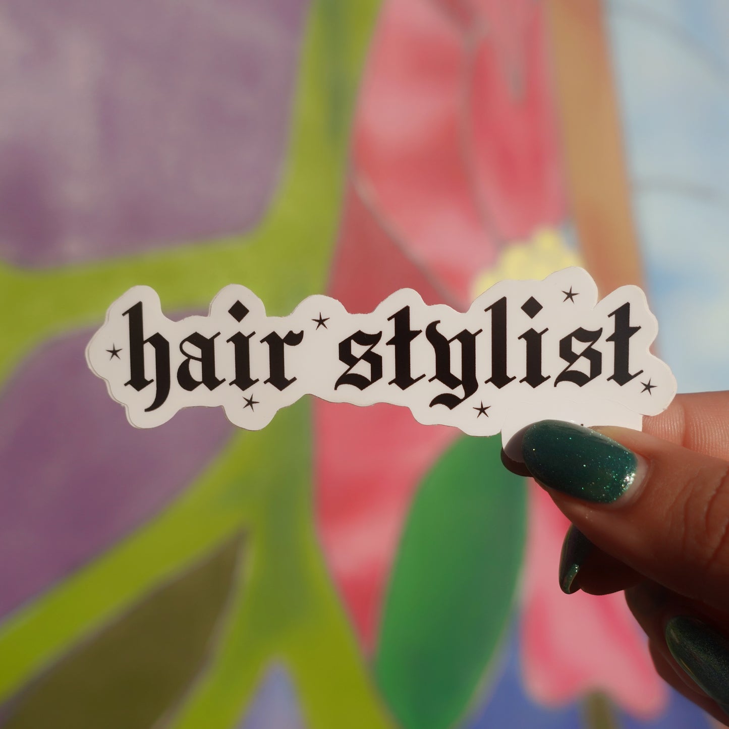 Sticker with 'hair stylist' text held by a hand against a colorful floral background