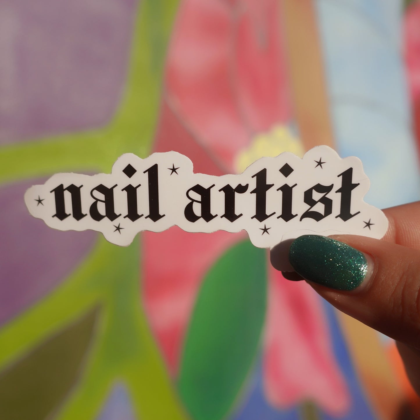 Sticker with 'nail artist' text held by a hand with green nail polish against a colorful background.