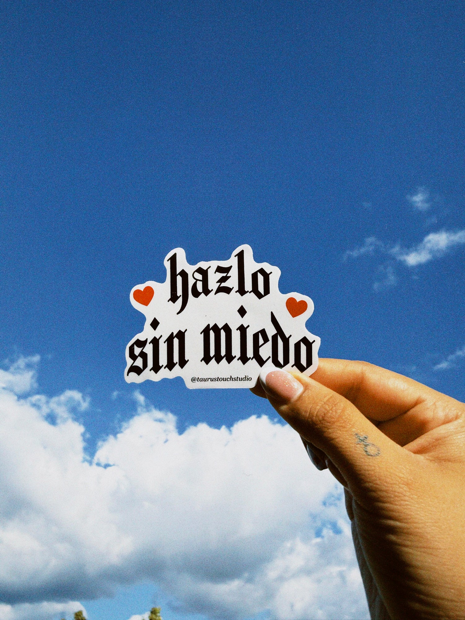 Hand holding a sticker with 'hazlo sin miedo' text against a blue sky.
