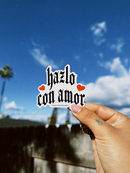 Hand holding a sticker with 'hazlo con amor' text against a blue sky with clouds and palm trees.